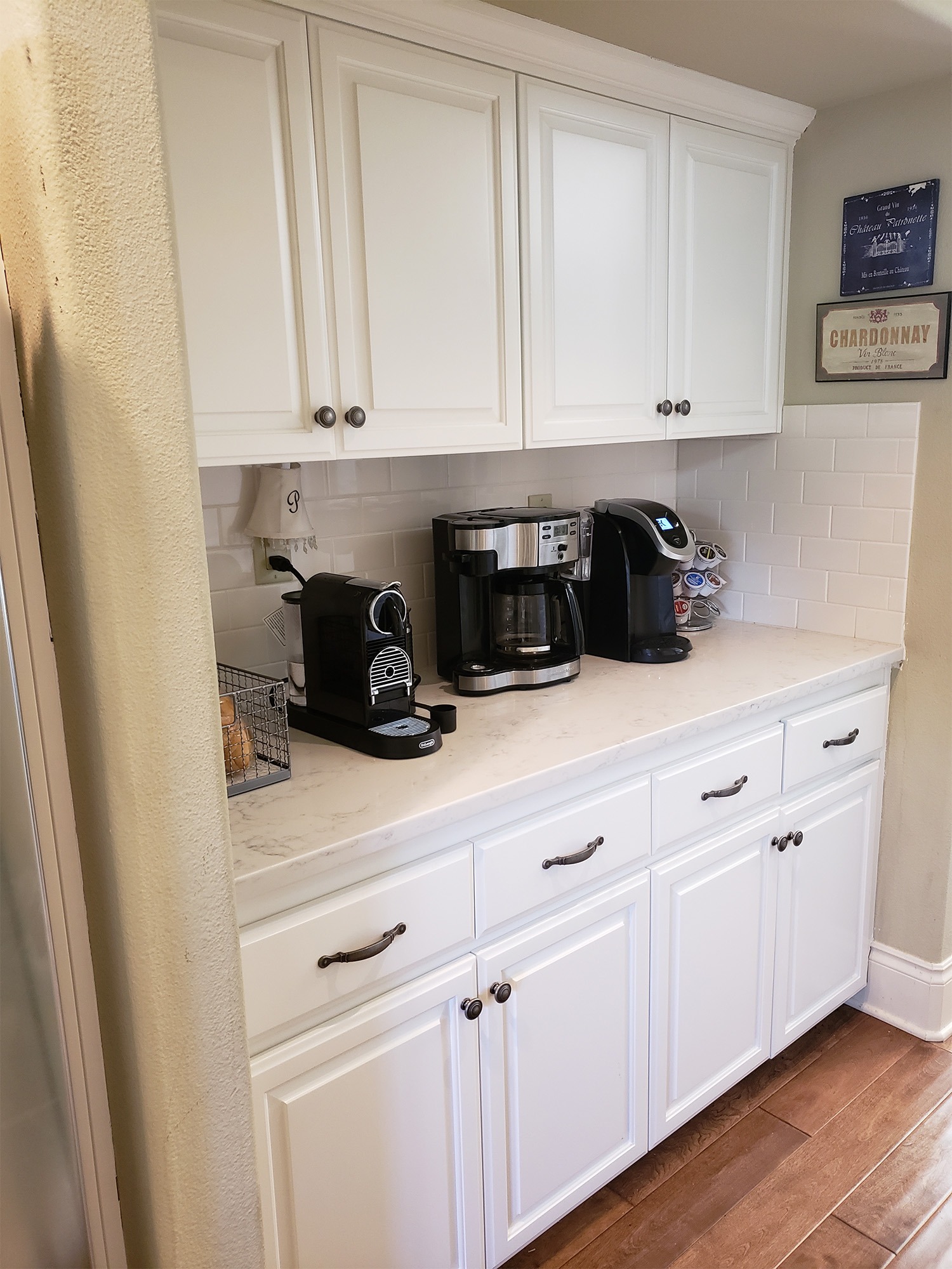 Kitchen coffee station featuring a white countertop with three coffee makers, storage basket, and decorative wall signs. Ideal for morning routines.
